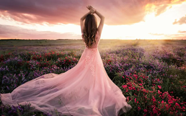 A woman in a flowing pink dress stands with her back to the camera in a vibrant flower field at sunset, creating a serene and dreamy mood in this HD desktop wallpaper.