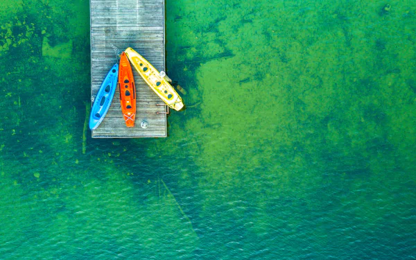 Aerial view of a wooden pier extending over clear green water with three colorful kayaks docked, captured in 4K Ultra HD resolution for a PC desktop wallpaper.
