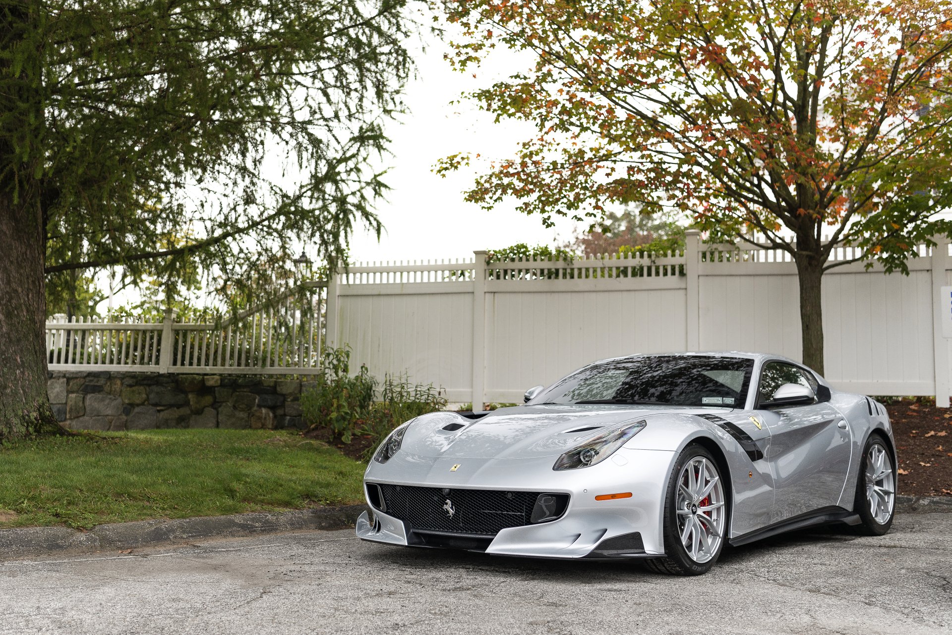 Silver Ferrari F12berlinetta supercar parked by a white fence and trees — 4K Ultra HD PC desktop wallpaper/background.