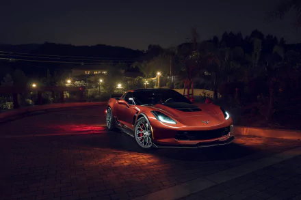 HD desktop wallpaper of an orange Chevrolet Corvette Z06 at night, parked on a lit driveway with headlights glowing against a dark suburban background.