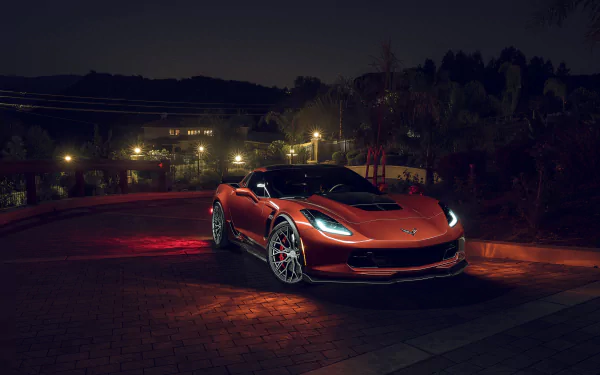 HD desktop wallpaper of an orange Chevrolet Corvette Z06 at night, parked on a lit driveway with headlights glowing against a dark suburban background.
