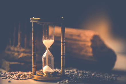 Close-up of a man-made brass hourglass with sand flowing against a warm, blurred backdrop — HD PC desktop wallpaper and background.