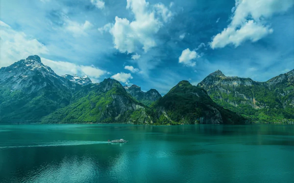 A serene lake surrounded by forested mountains under a partly cloudy sky, with a small boat moving across the turquoise water. HD wallpaper and background.
