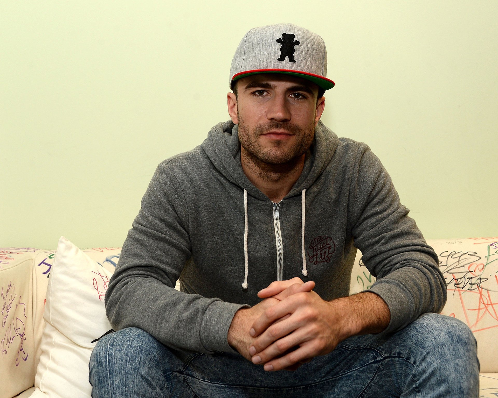 HD desktop wallpaper featuring a man in a grey hoodie and baseball cap, seated and looking at the camera.