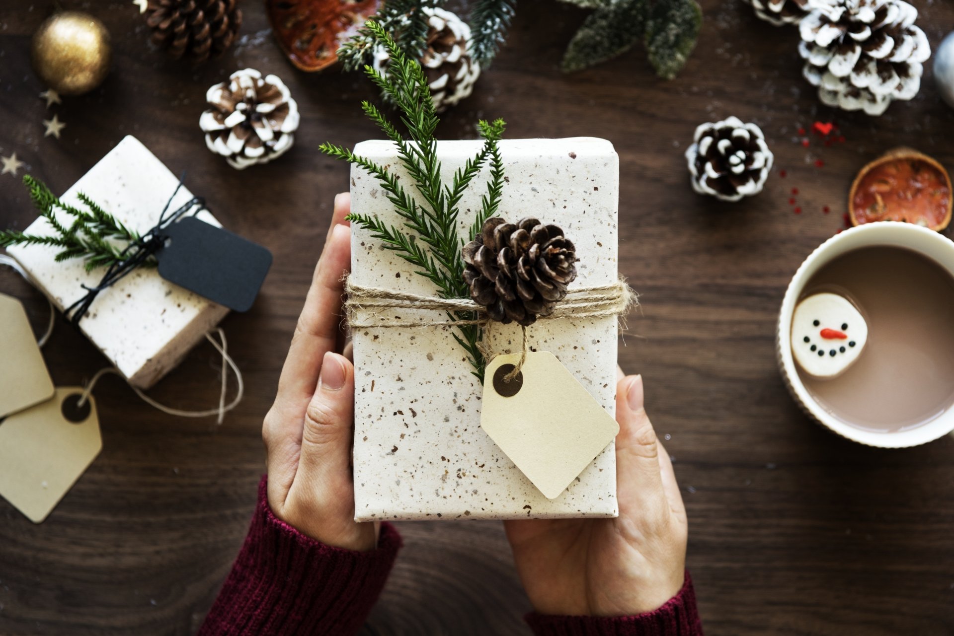 A hand holds a gift wrapped in speckled white paper, adorned with a pine cone and greenery, set against a rustic wooden table with more gifts and a cup of hot chocolate.