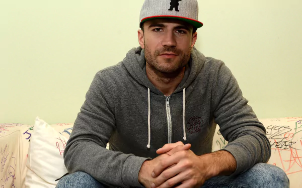 HD desktop wallpaper featuring a man in a grey hoodie and baseball cap, seated and looking at the camera.