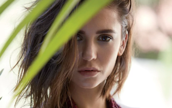Close-up HD portrait of Canadian actress Nina Dobrev with brown eyes and brunette hair, framed by blurred greenery in the foreground and soft background.