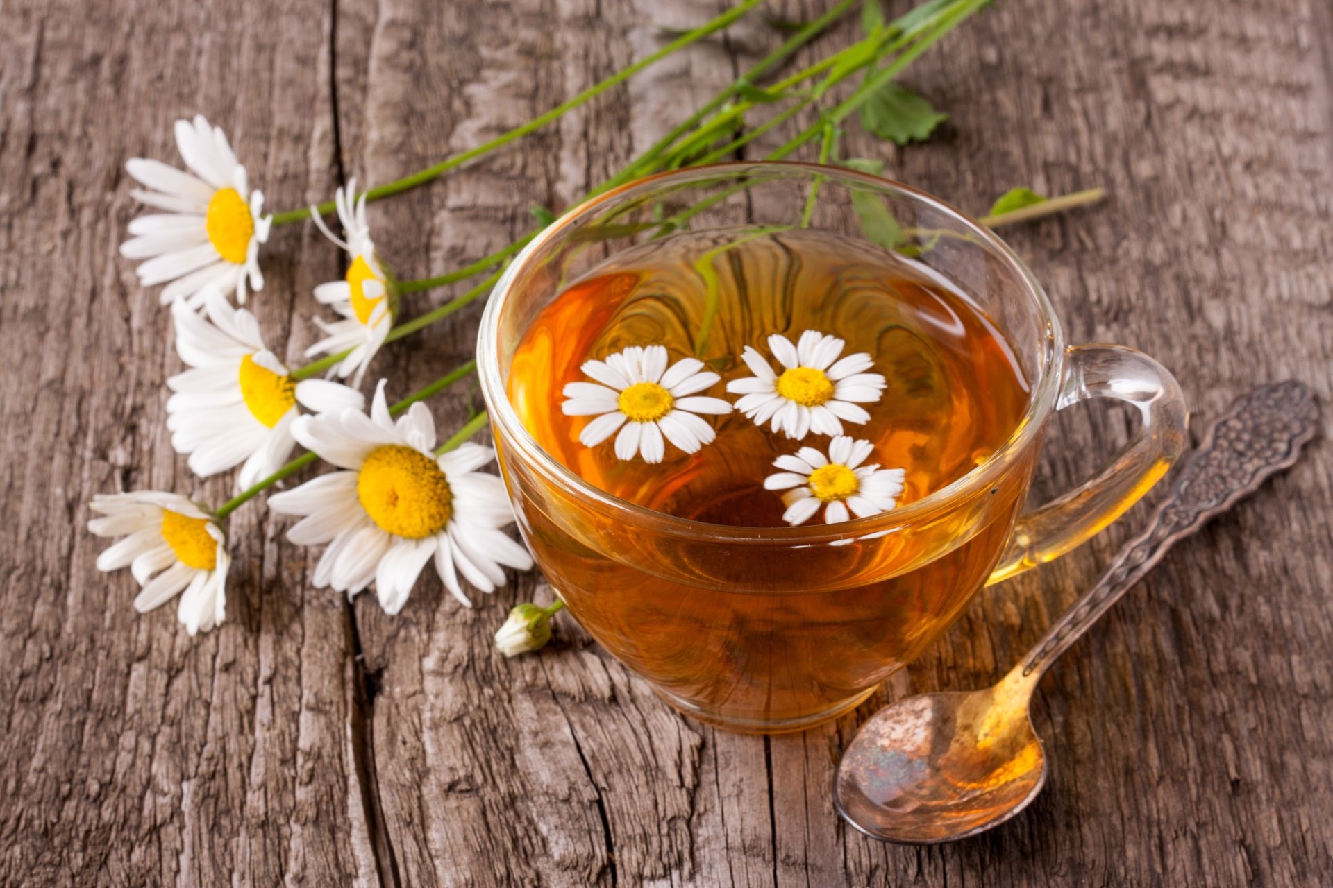 4K Ultra HD image of a clear cup of chamomile tea with floating white chamomile flowers, resting on a rustic wooden surface alongside fresh chamomile blooms and a spoon.