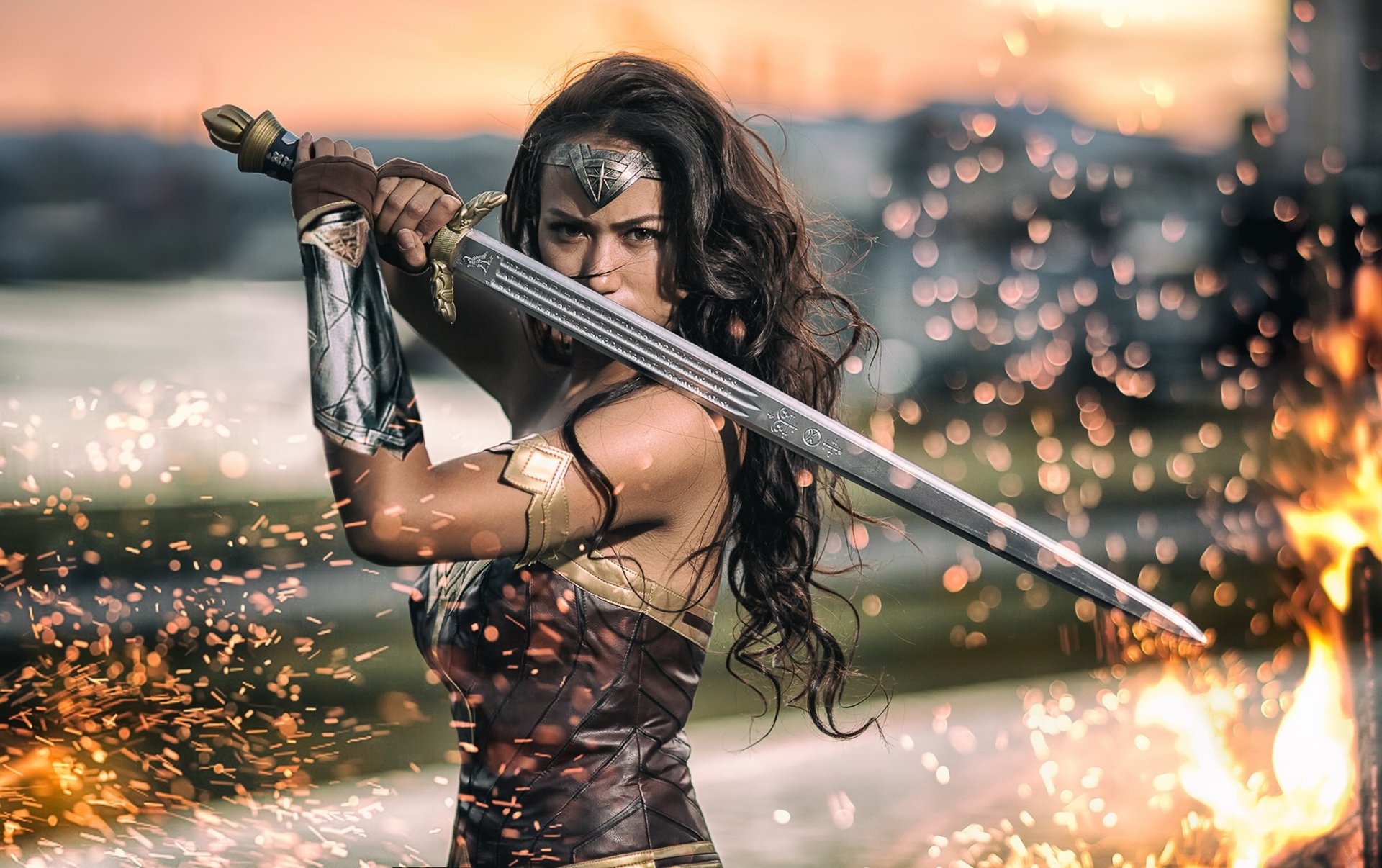 A model cosplaying as Wonder Woman holds a sword in a bokeh-filled, fiery background, featuring a brunette with a determined expression. The HD desktop wallpaper emphasizes depth of field and dynamic action.