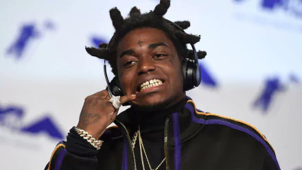 HD desktop wallpaper featuring a smiling person with distinctive hair, gold teeth, and headphones, tagged with Kodak Black, on a blurred background.