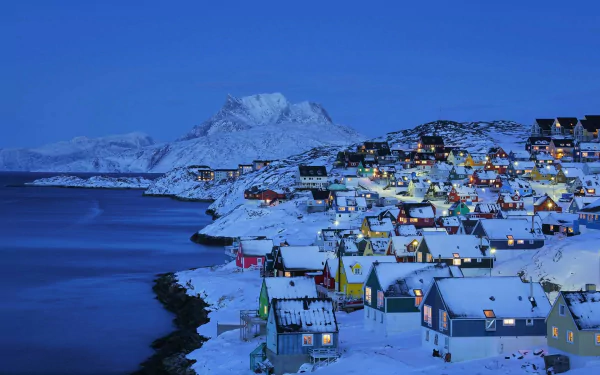 HD desktop wallpaper of a snowy winter scene in a Norwegian village in Lofoten, with houses illuminated under a wintry blue sky, mountains, and calm sea in the background.