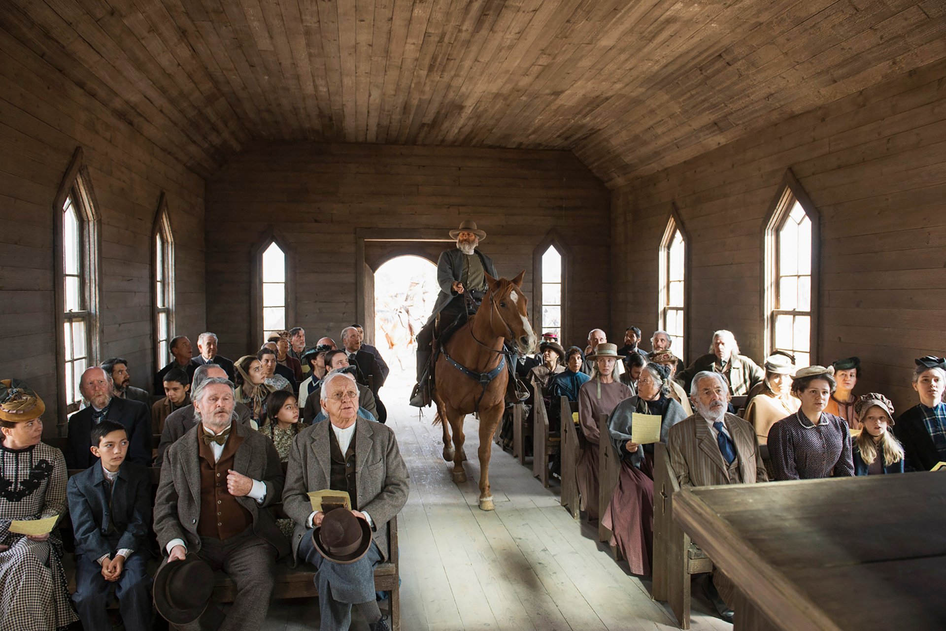 HD desktop wallpaper from Godless featuring a person on horseback inside a wooden building surrounded by a crowd of onlookers.