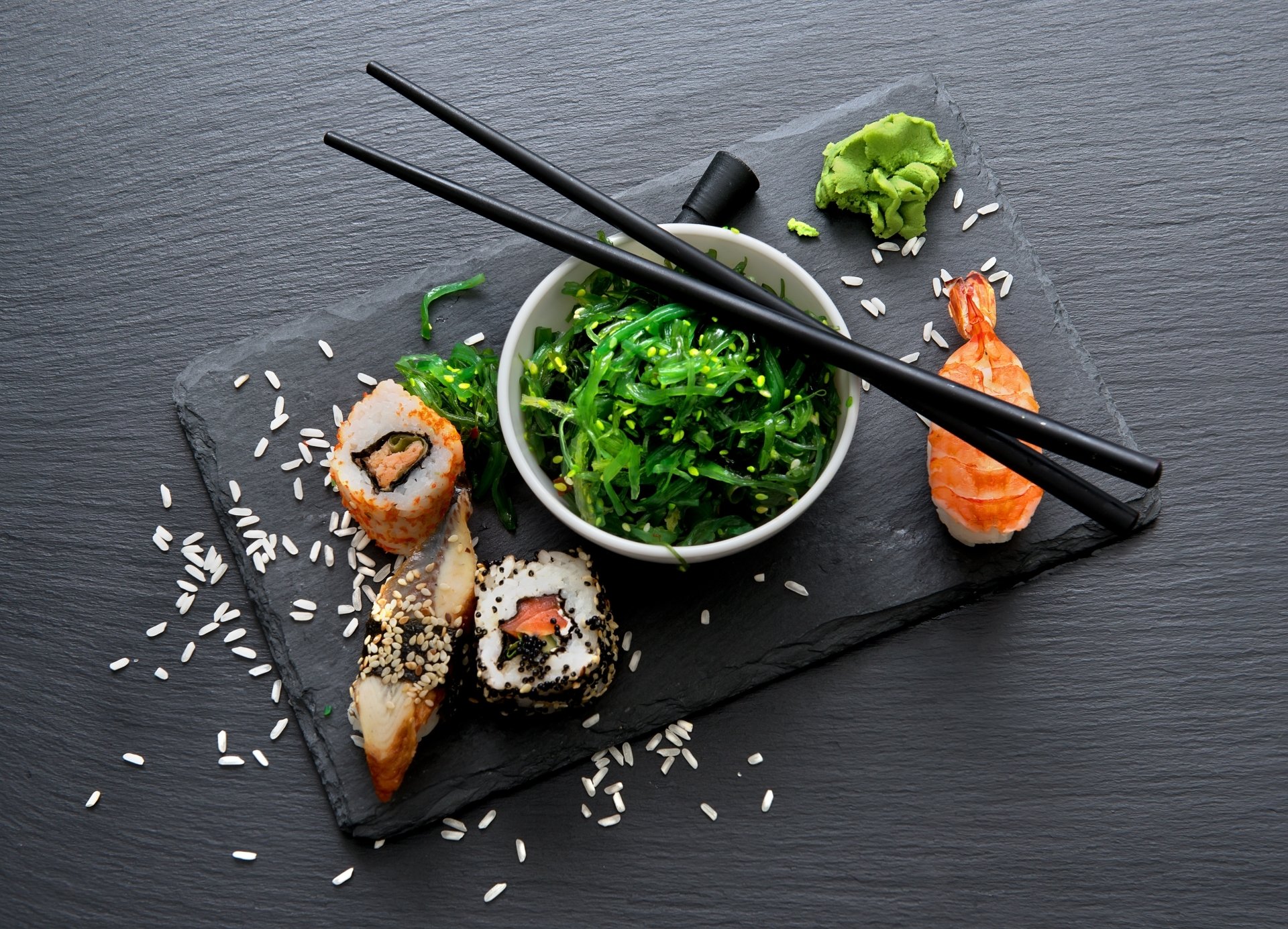 2K Quad HD desktop wallpaper still life of sushi and seafood: maki rolls, shrimp nigiri, seaweed salad on a slate board with scattered rice and crossed black chopsticks.
