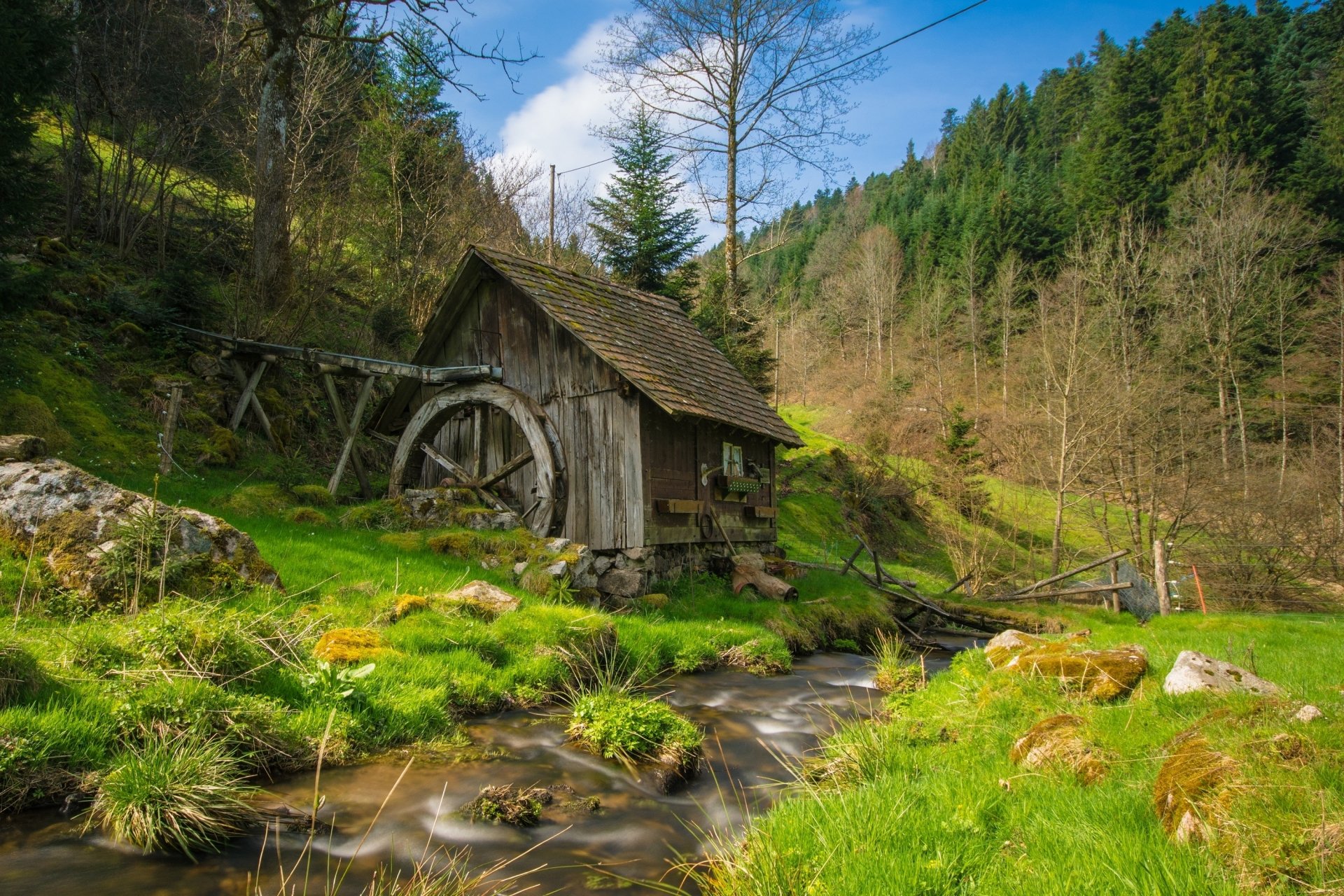 HD desktop wallpaper featuring a serene scene with a rustic watermill by a stream, surrounded by green grass and trees on a hillside.