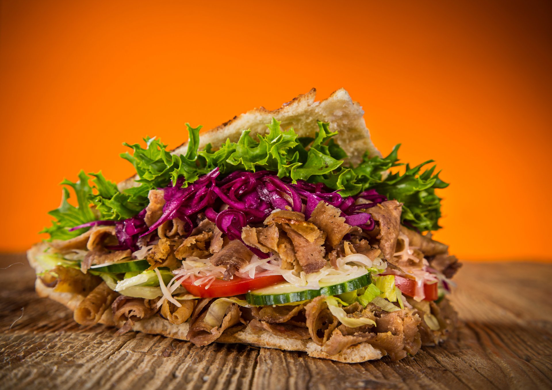 5K Ultra HD PC desktop wallpaper: close-up stacked sandwich with sliced meat, shredded purple cabbage, lettuce, tomato and cucumber on rustic wood against an orange backdrop.
