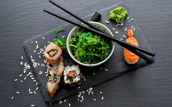 2K Quad HD desktop wallpaper still life of sushi and seafood: maki rolls, shrimp nigiri, seaweed salad on a slate board with scattered rice and crossed black chopsticks.