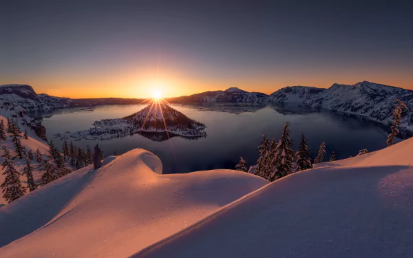 Snow-covered landscape at Crater Lake, Oregon, during winter sunrise. The serene lake is surrounded by a snowy island and mountain scenery, capturing the beauty of nature in this HD wallpaper background.