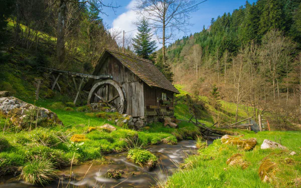 HD desktop wallpaper featuring a serene scene with a rustic watermill by a stream, surrounded by green grass and trees on a hillside.