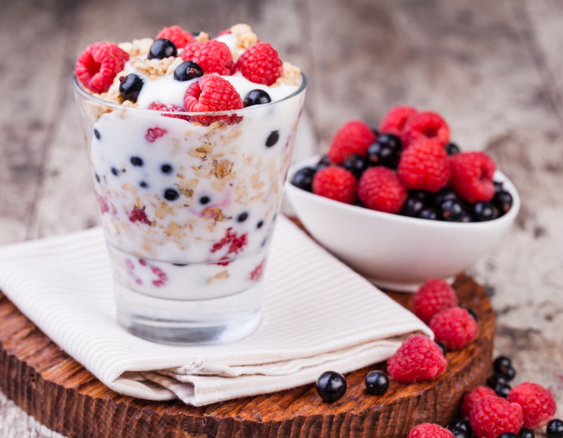 Close-up still life of a glass filled with muesli, milk, raspberries, and blueberries beside a bowl of mixed berries on a wooden surface, captured in 4K Ultra HD quality.