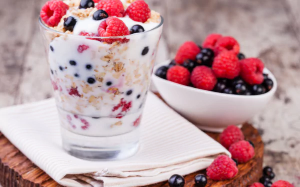 Close-up still life of a glass filled with muesli, milk, raspberries, and blueberries beside a bowl of mixed berries on a wooden surface, captured in 4K Ultra HD quality.