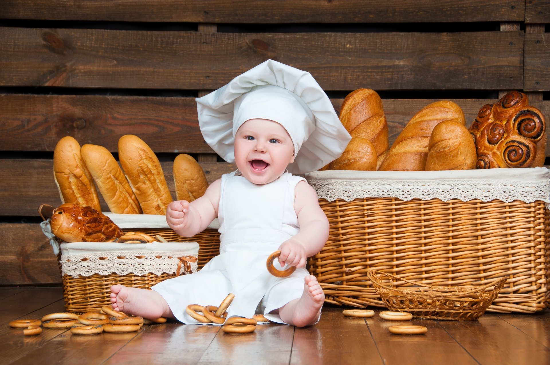 A smiling baby dressed as a chef sits on a wooden floor surrounded by baskets of bread and pretzels in a 4K Ultra HD desktop wallpaper.