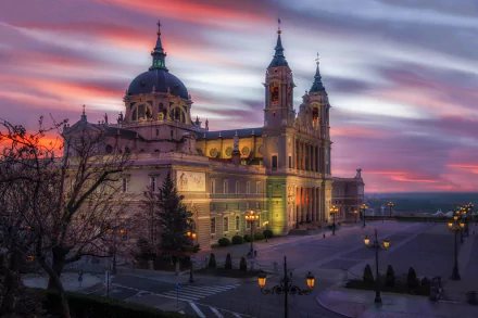 Almudena Cathedral in Madrid illuminated at dusk, with vibrant sunset colors and dramatic lighting highlighting its architectural details in a serene square setting.