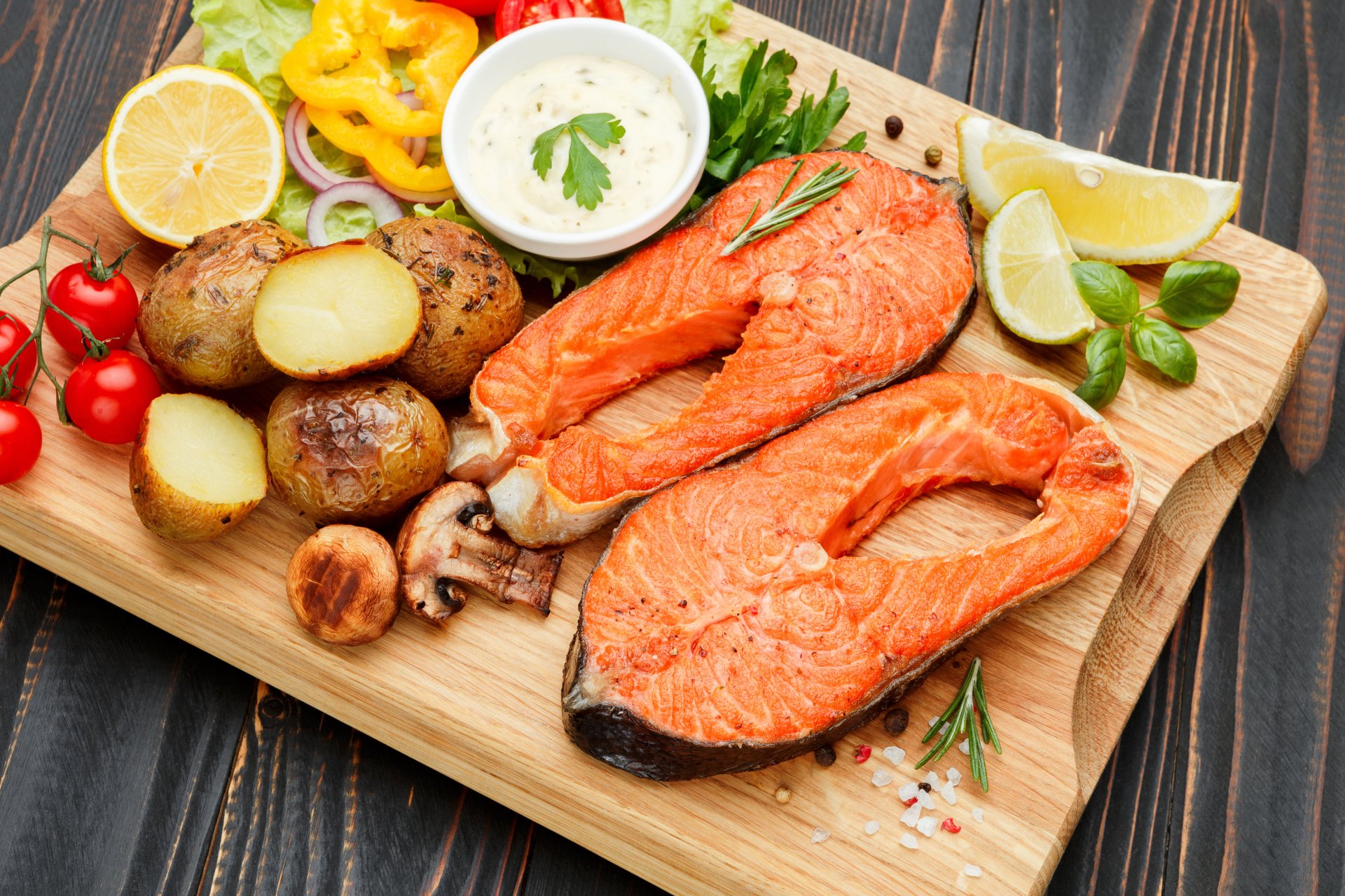 A vibrant 4K Ultra HD still life of cooked salmon steaks with roasted potatoes, cherry tomatoes, lemon slices, and dipping sauce on a wooden board, showcasing seafood and food.