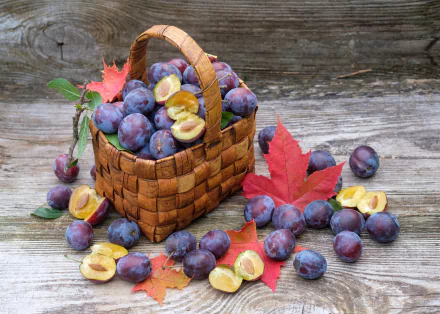 A woven basket filled with ripe plums, surrounded by halved plums and autumn leaves on rustic wooden surface, presented as an HD PC desktop wallpaper still life.