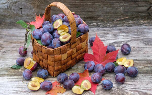 A woven basket filled with ripe plums, surrounded by halved plums and autumn leaves on rustic wooden surface, presented as an HD PC desktop wallpaper still life.