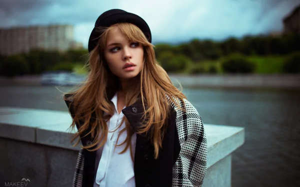 A portrait of a woman with long hair wearing a hat, taken outdoors with a blurred background featuring water and green trees. The image is tagged with Anastasiya Scheglova and has a depth of field effect.