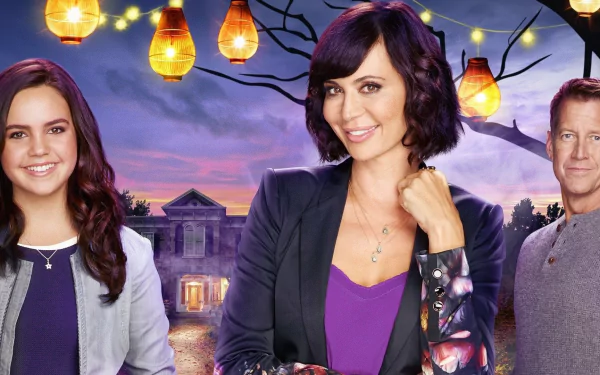 HD PC desktop wallpaper background of the TV show Good Witch: three characters smiling in front of a lit Victorian house at dusk with hanging lanterns and a purple sky.
