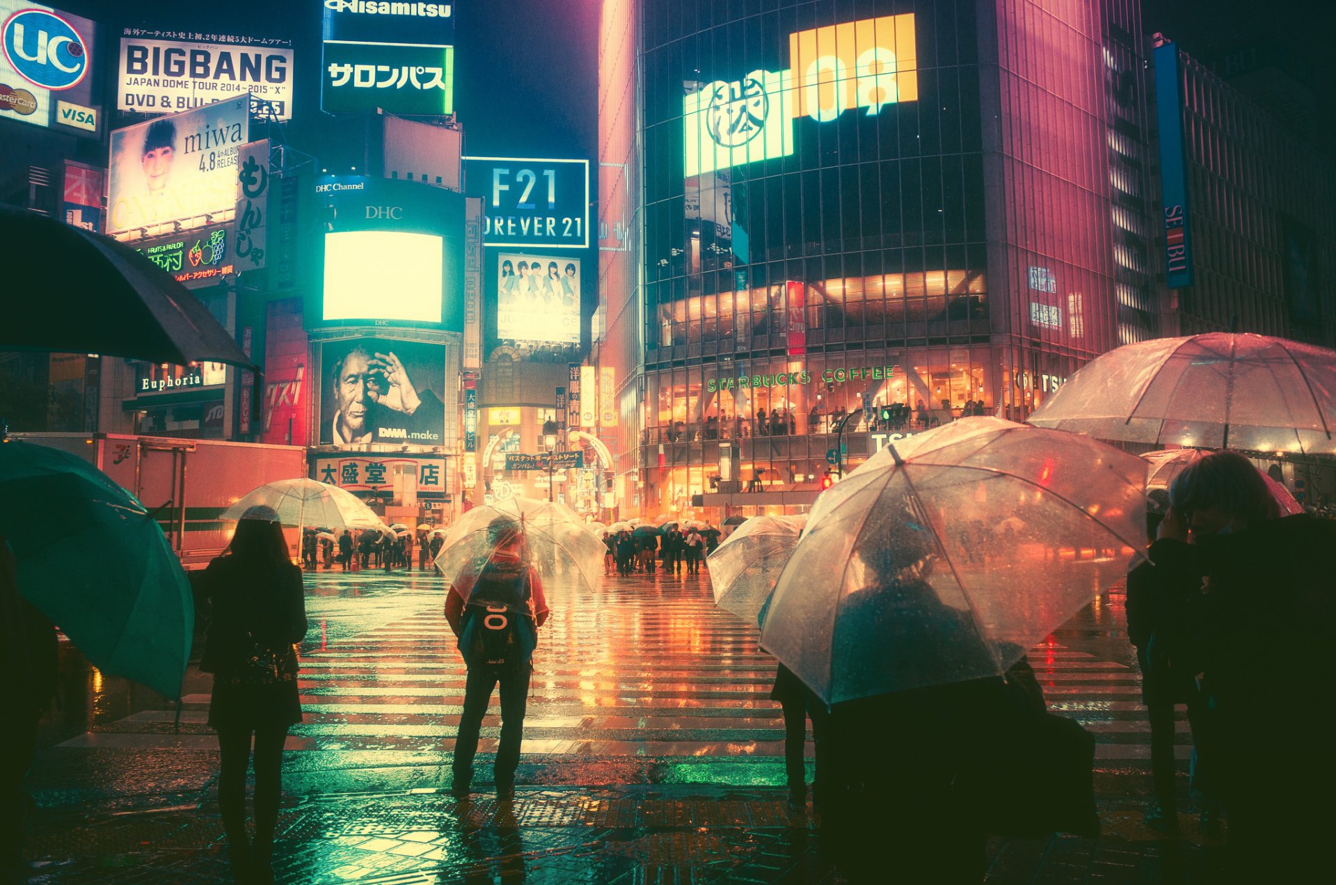 A nighttime city scene in Japan with glowing neon billboards and people holding umbrellas crossing a wet street, captured in HD for a vibrant desktop wallpaper.