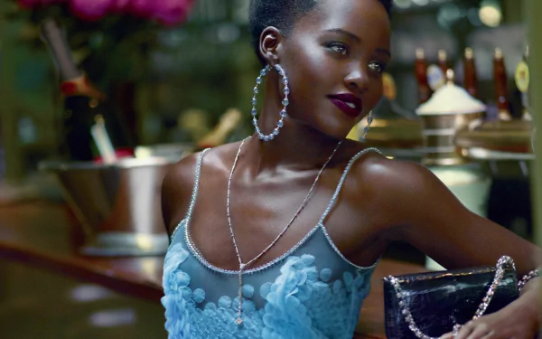 HD PC desktop wallpaper featuring celebrity Lupita Nyong'o in a stylish blue dress with large hoop earrings, posing confidently in a dimly lit setting.