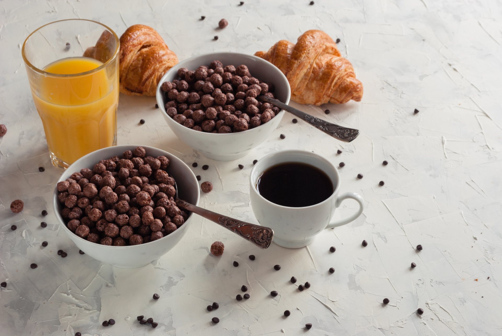 4K Ultra HD still life of a breakfast spread featuring two bowls of chocolate cereal, a cup of coffee, a glass of orange juice, and two croissants on a white surface.