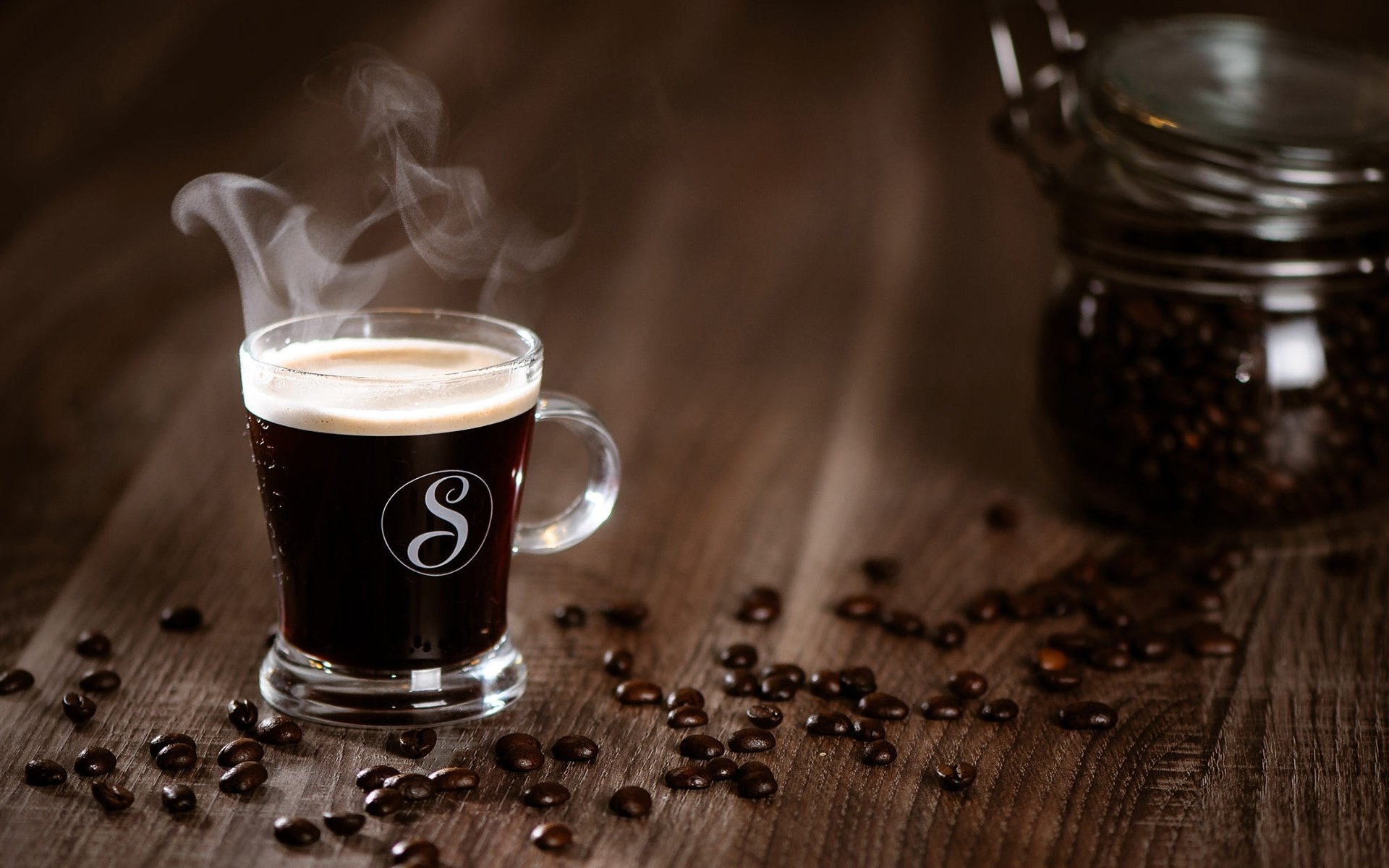 HD desktop wallpaper showing a steaming cup of coffee with scattered coffee beans and a glass jar on a wooden surface.