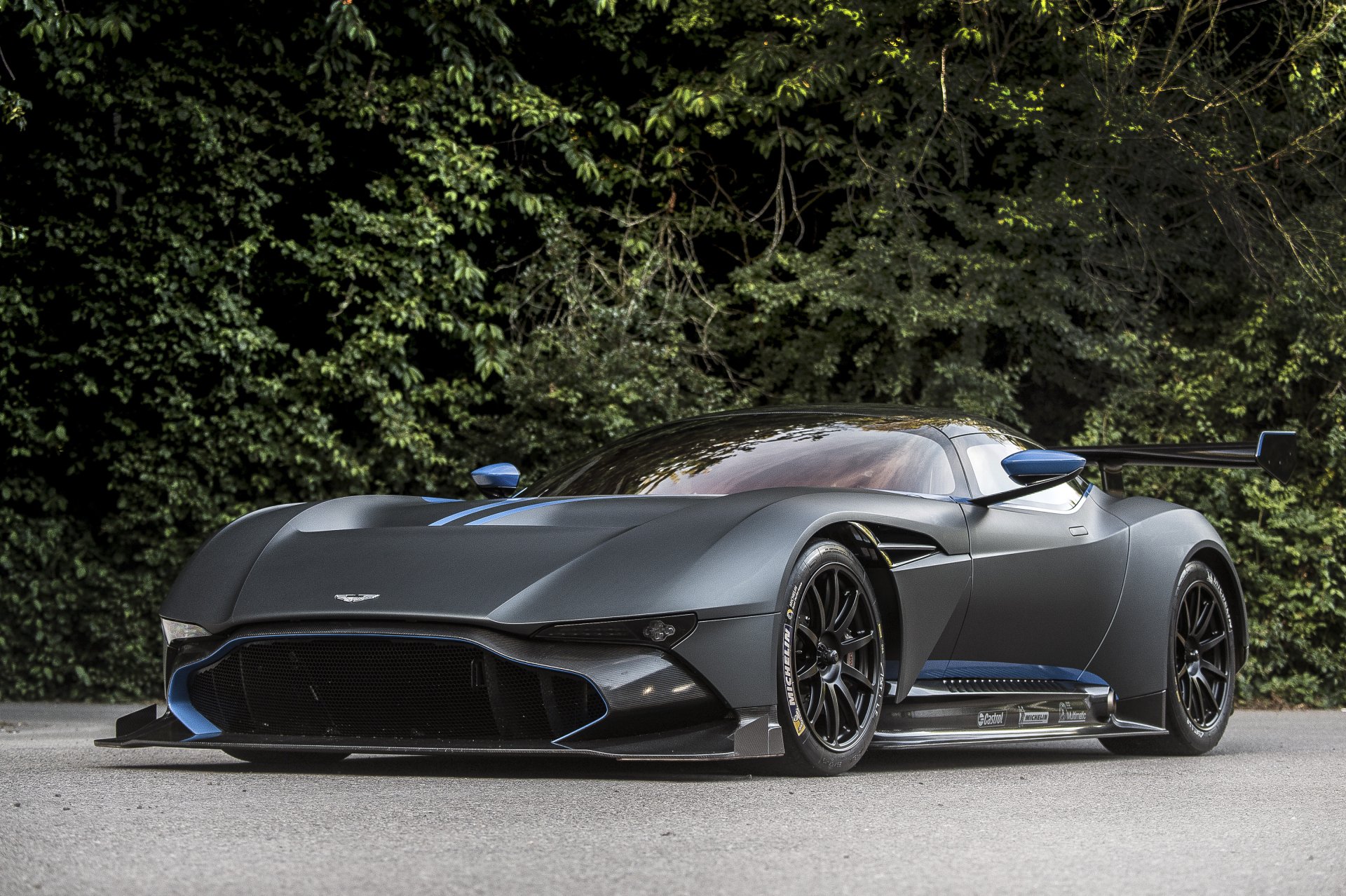 A sleek, dark gray Aston Martin Vulcan supercar parked on a road with dense green foliage in the background, captured in 4K Ultra HD for a PC desktop wallpaper.