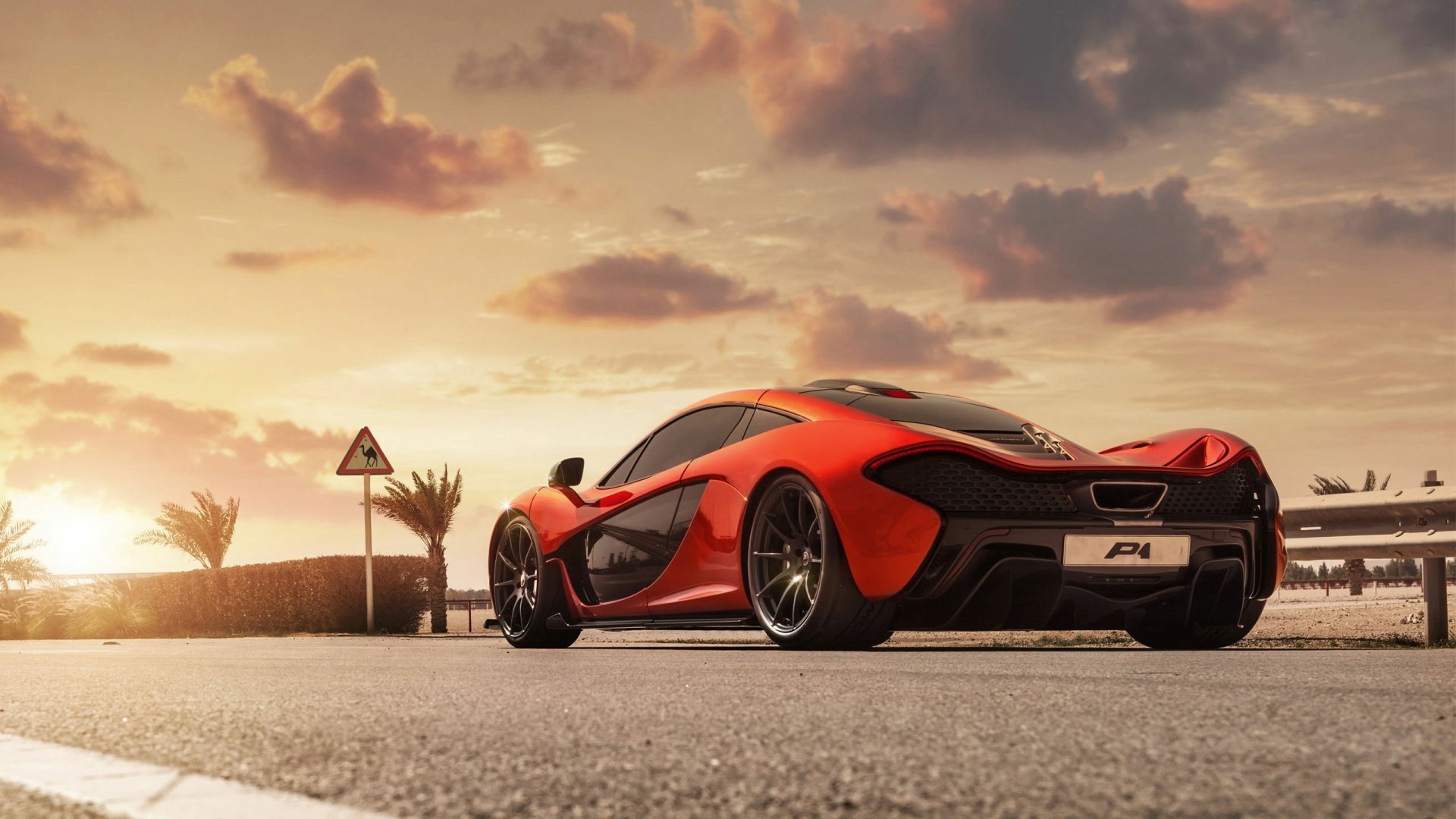 HD desktop wallpaper featuring an orange McLaren P1 supercar parked on a sunlit road under a dramatic sky.