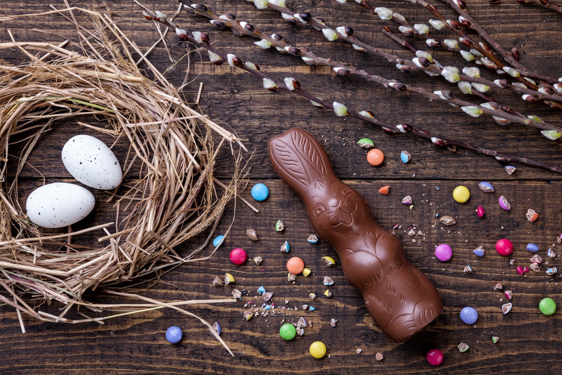 A chocolate Easter bunny, colorful candy pieces, and a nest with white speckled eggs arranged on a rustic wooden surface, creating a festive holiday still life in 4K Ultra HD.
