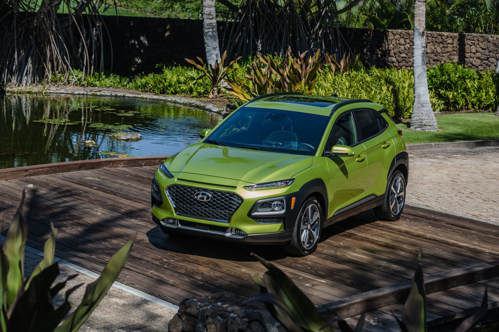 Green Hyundai Kona SUV parked near a pond with tropical plants in the background, shown as an HD PC desktop wallpaper and background.