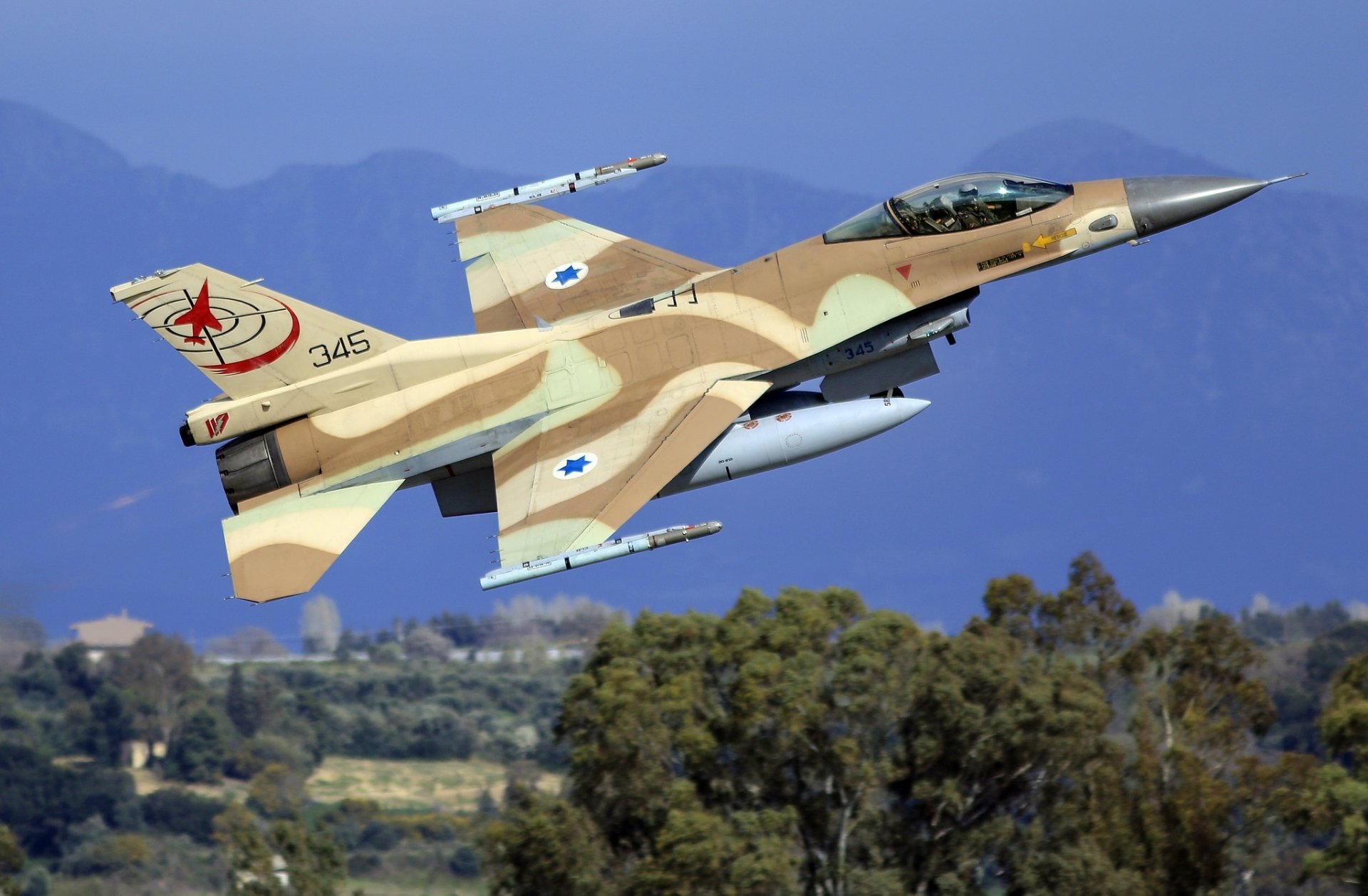 A General Dynamics F-16 Fighting Falcon jet fighter in desert camouflage flies over a landscape with trees and mountains in the background.