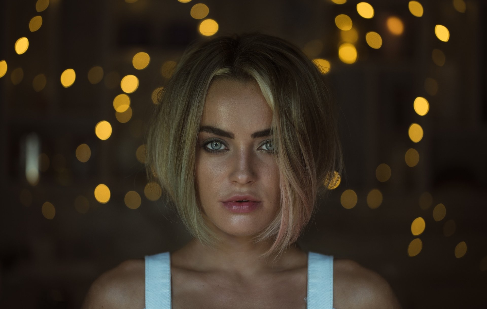 Close-up of a blonde woman with short hair and striking blue eyes against a dark background with warm bokeh lights, captured in HD for a desktop wallpaper.