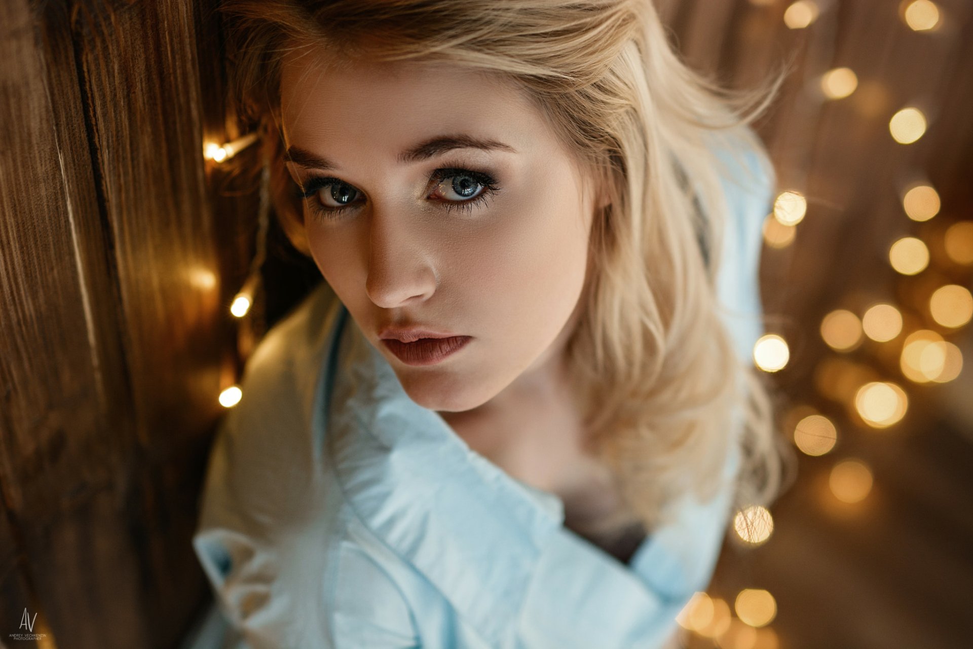 HD desktop wallpaper featuring a close-up of a blonde woman with blue eyes, softly illuminated by warm bokeh lights in the background.