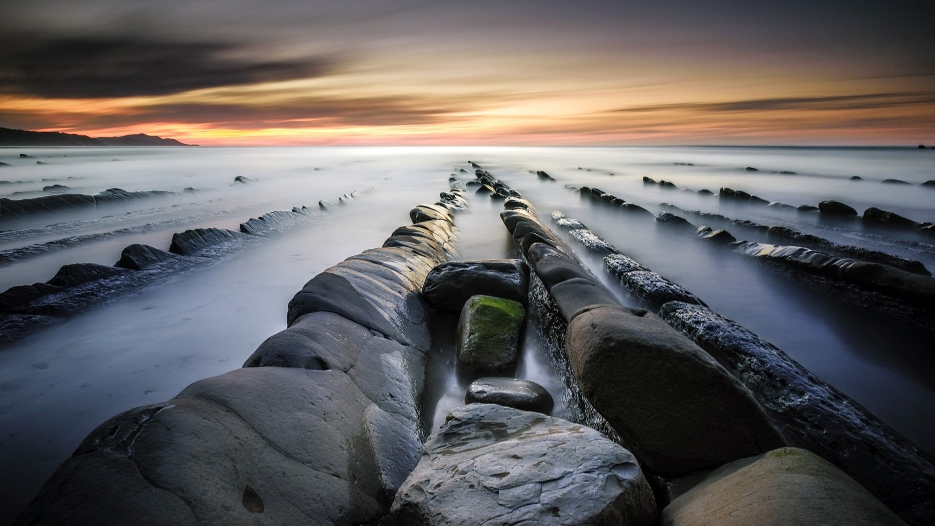 HD PC desktop wallpaper and background: nature seascape of the ocean at sunset, smooth long-exposure waves flowing between jagged rock ridges toward the horizon.