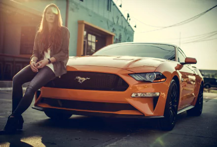 A redhead woman with long hair, wearing jeans and a jacket, leans against an orange Ford Mustang. The scene is set in an urban environment, captured as a HD desktop wallpaper and background.