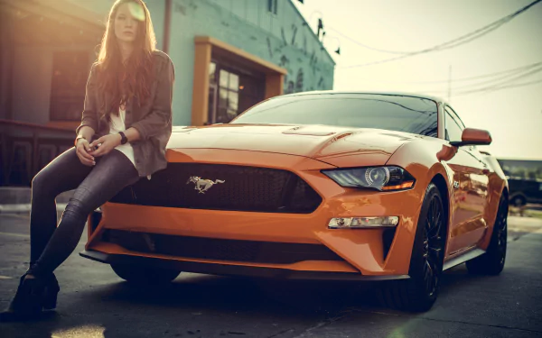 A redhead woman with long hair, wearing jeans and a jacket, leans against an orange Ford Mustang. The scene is set in an urban environment, captured as a HD desktop wallpaper and background.