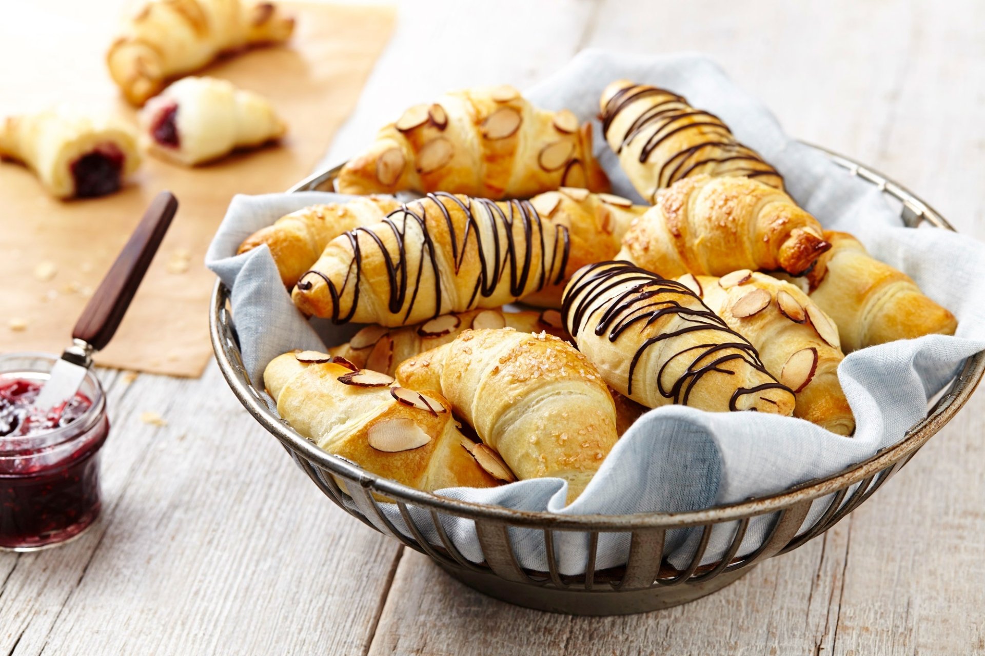 HD desktop wallpaper featuring a basket of golden croissants drizzled with chocolate, surrounded by jam and sliced almonds, capturing a delicious Viennoiserie breakfast scene.
