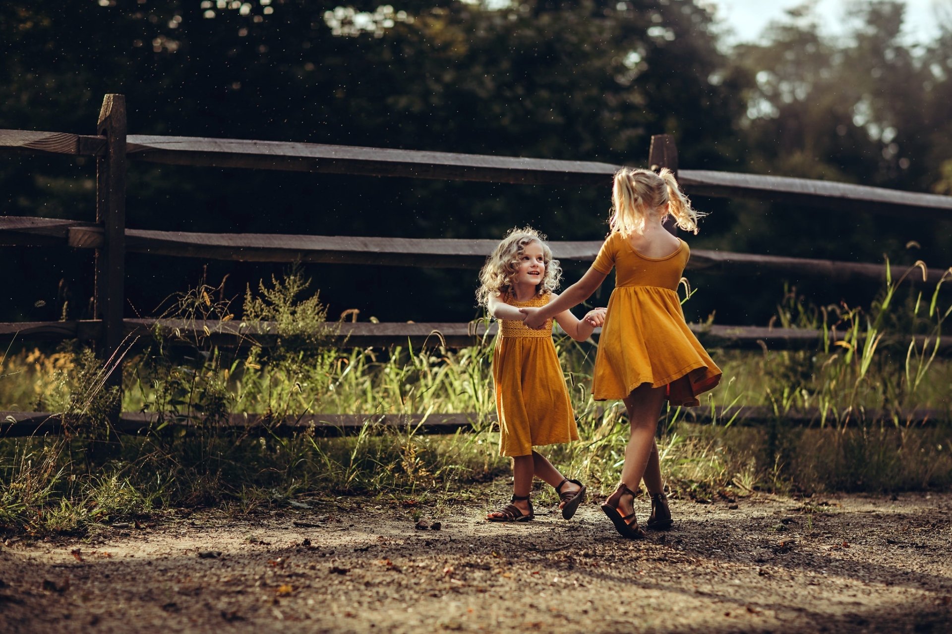 Two blonde little girls in orange dresses hold hands and play outdoors near a wooden fence, captured in a vibrant HD photography wallpaper background.