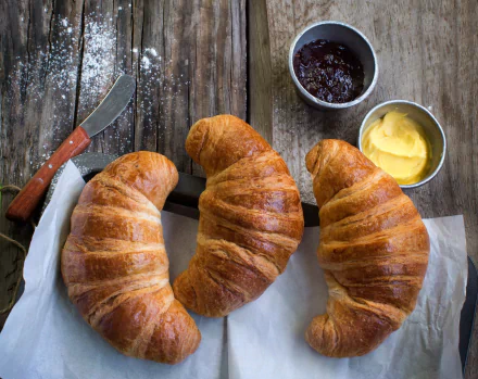 HD PC desktop wallpaper of three golden croissants on parchment with butter and jam on a rustic wooden table — breakfast viennoiserie food background