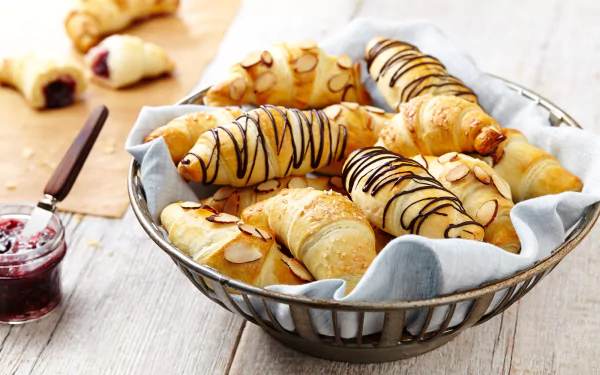 HD desktop wallpaper featuring a basket of golden croissants drizzled with chocolate, surrounded by jam and sliced almonds, capturing a delicious Viennoiserie breakfast scene.