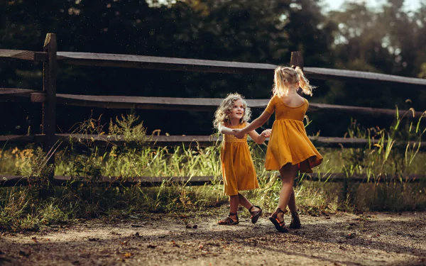 Two blonde little girls in orange dresses hold hands and play outdoors near a wooden fence, captured in a vibrant HD photography wallpaper background.